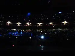 A darkened concert venue. People are in multiple levels of seating, and tiny lights are scattered throughout the audience. On the left, performers stand on a stage lit with blue lighting.