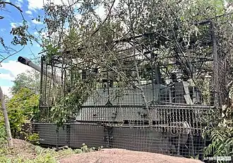Ukrainian M109A4 with anti-drone metal cage surrounded by trees in daytime