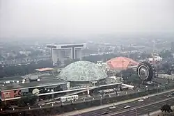 In a misty aerial view, a large building, a major road, a helipad, and two colored domes are visible.