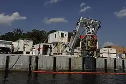 A dockside cluttered with containerised units and a gantry, which supports a bell over the water.