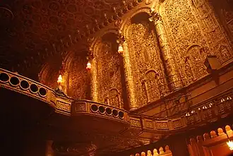 The interior of the auditorium's left wall, looking from the orchestra level toward the balcony. The wall contains elaborate carvings and engaged columns, and the balcony has a carved balustrade.