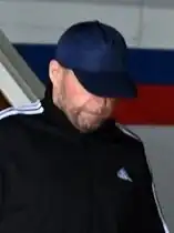A middle-aged male with their head pointing down, with much of it covered by a blue ball cap. He also displays a partial beard, and is wearing a black adidas jacket