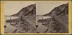 Two men standing on the Hudson River shore with railroad tracks paralleling the shoreline and a mountain off in the distance