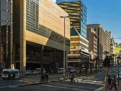 A busy, divided urban street seen from its right side, looking uphill to blue sky and a green hill in the distance. Tall buildings line the far side; the one nearest the viewer is too high to be contained within the image. Near the viewer is an intersection with a traffic light, with pedestrians crossing and waiting to cross