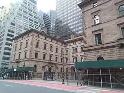 The courtyard and the houses' brownstone facades as seen from across Madison Avenue in early 2021. There are green sidewalk sheds in front of the houses.