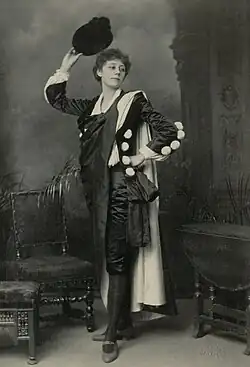 Young woman with short curly hair standing, posed in flamboyant male clothing, with hat and cape in indoor setting