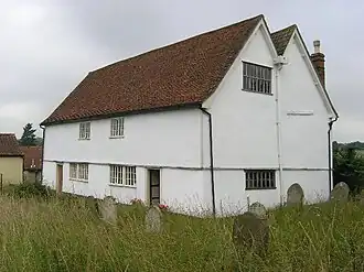 A white building with a red-tied roof seen from an angle and looking more like a house than a chapel. It has two storeys, a double gable, two windows on the end and on the entrance front are two doors and two windows in each storey.