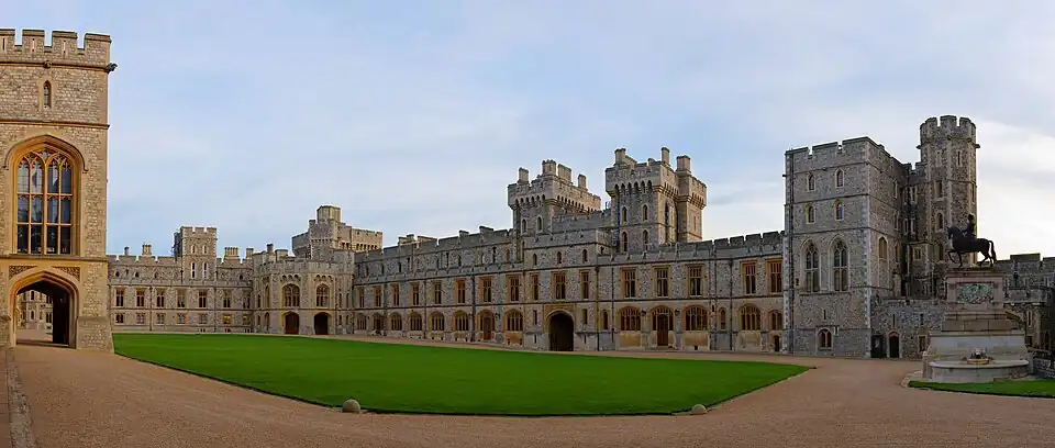 A photograph of a grey Gothic quadrangle with a green grass square in the middle. On the left, a block of the building makes up the near ground. A gatehouse is in the middle of the right-hand part of the quadrangle.