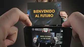 A person is holding a sign reads "BIENVENIDO AL FUTURO". Another person is holding an iPhone to translate the sign. The phone shows "WELCOME TO THE FUTURE", and "Spanish to English".