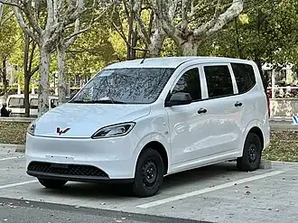 Third generation Wuling Hongguang EV front quarter view in white paint. It is parked in a parking lot rear-in outdoors on a sunny evening with trees in the background.