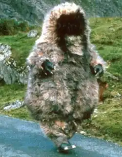 A large, fluffy, grey-ish brown costume, resembling a bipedal furry beast with no discernible face, walking down a path in a grassy, mountainous area.