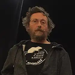 A man with glasses and a beard, wearing a black hoodie over a T-shirt with French text and a rat illustration, standing in front of a dark background.
