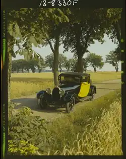 A historical image from 1967 of a black Bugatti Type 40 coupe with bright yellow doors. The car is parked next to the road, surrounded by fields of barley and trees. There is no driver in the car.